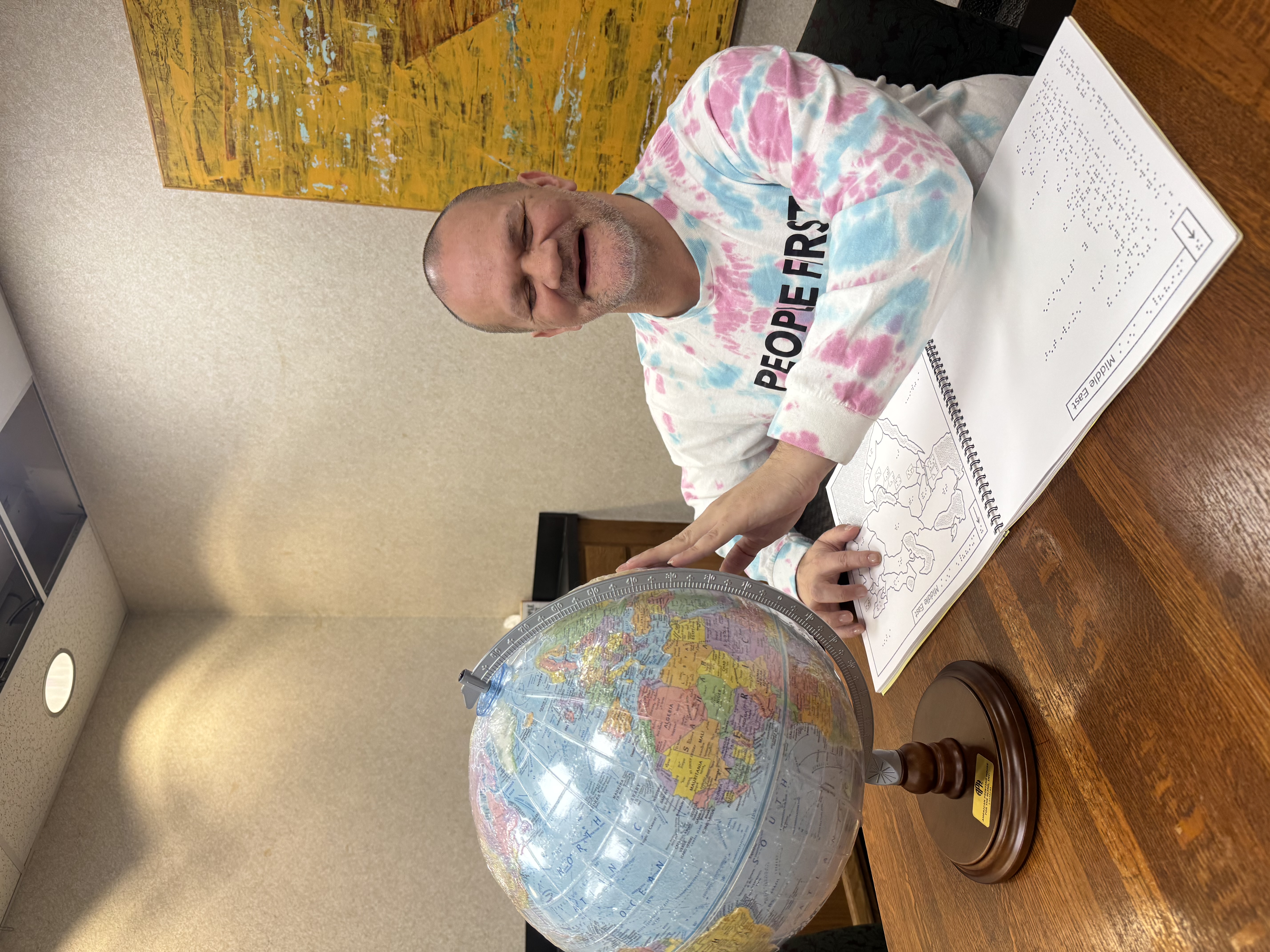 Dale smiles as he has one hand feeling a spot on the braille globe, and one on a map book in front of him.