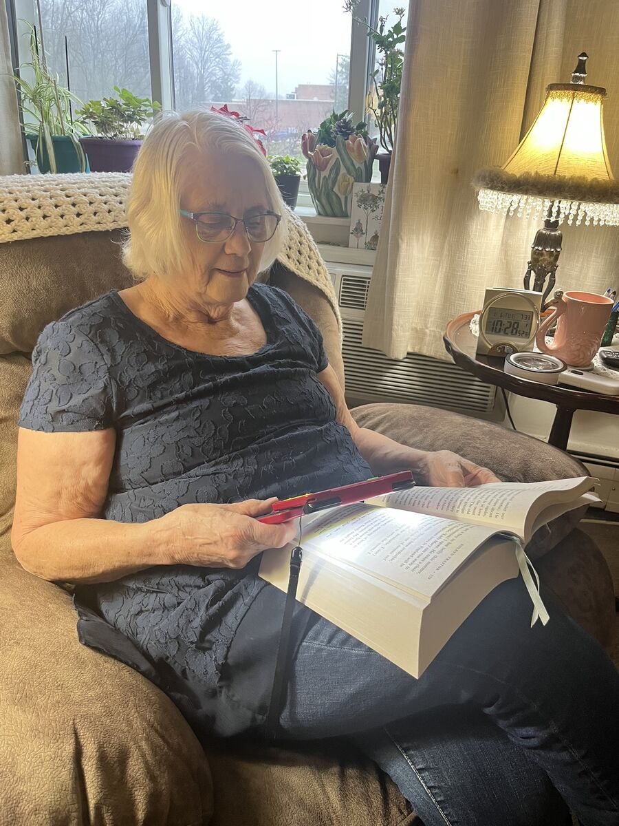 Carrie uses an electronic magnifier to read her large print book while sitting in a comfy lounge chair in her living room. 