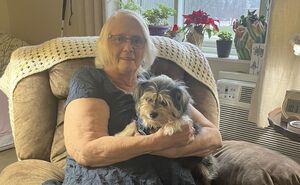 Carrie smiles for a photo while holding her small dog and sitting on a comfy chair in her living room.