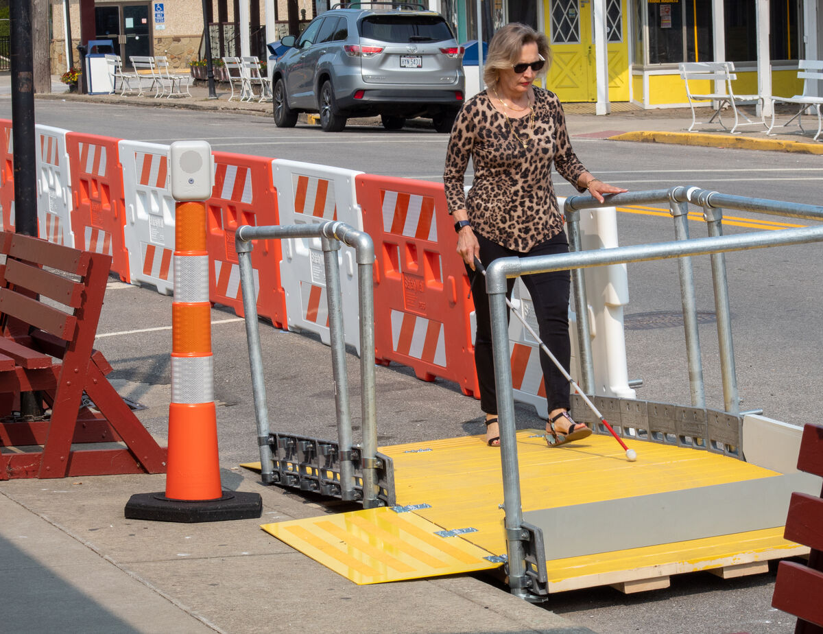 A woman in a cheetah print shirt uses her white cane to navigate a street with construction  partitions.