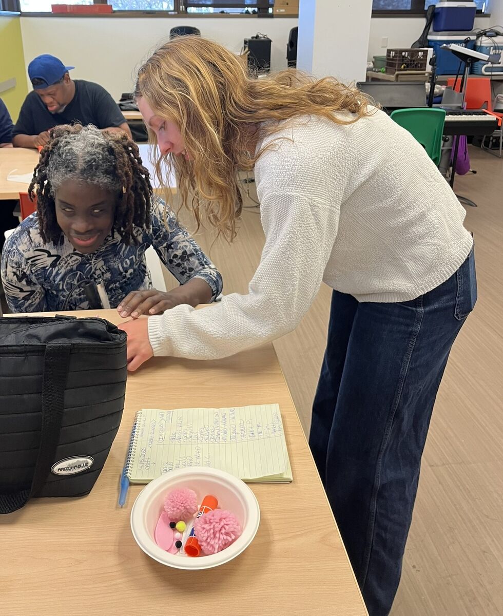 Lily leans over to help a woman participating in arts and crafts. Behind them, a man sit on a table working on his craft.