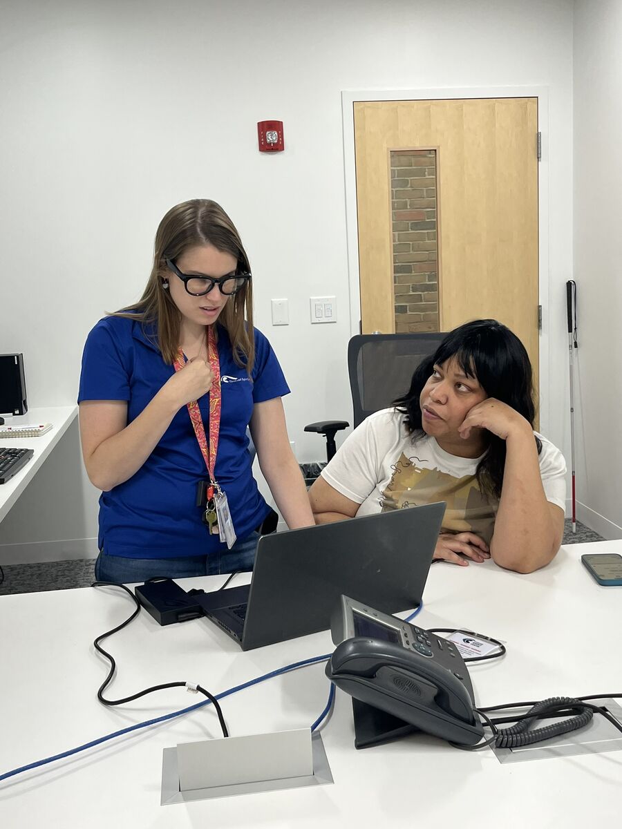 Chacity sits at a desk as her computer training instructor stands next to her and explains how to do something on her laptop. Chacity is looking up at her instructor.