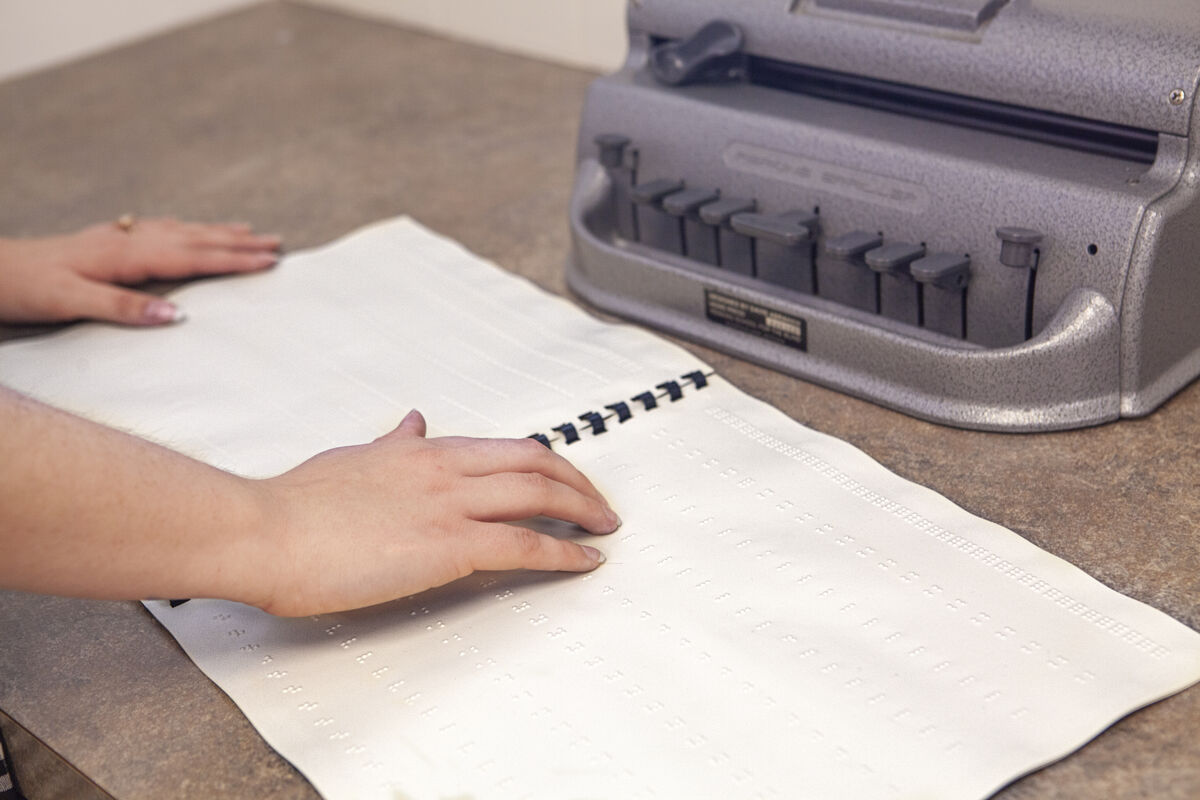 A hand moves across a book of braille. A Perkins Brailler is in the background.