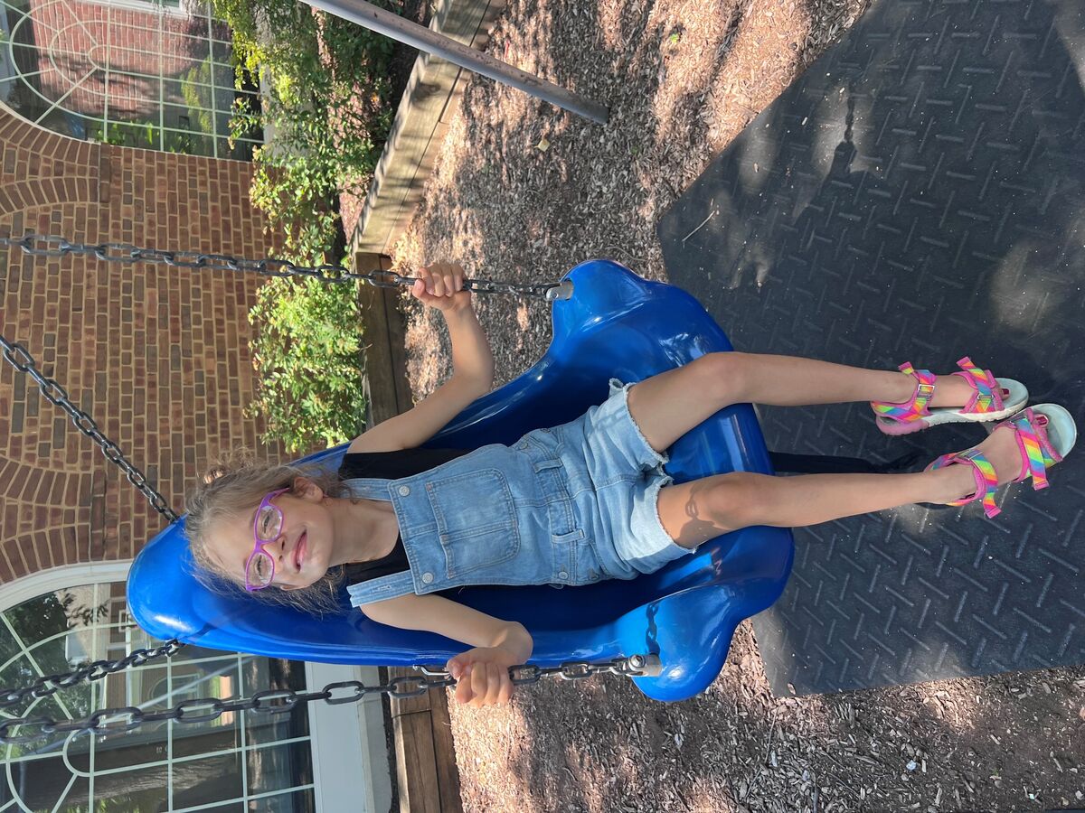 Kaylee sits in an accessible swing on a playground, and is smiling while wearing her new purple glasses.