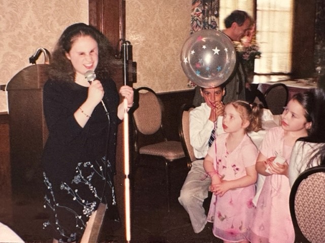 Nicole speaks into a microphone while holding her white cane at a party. 
