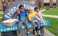 Three preschoolers sit on a bench at CSC's playground while holding on to each other and smiling at the camera.