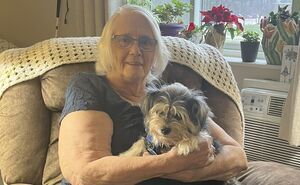 Carrie smiles for a photo while holding her dog in a comfy arm chair.
