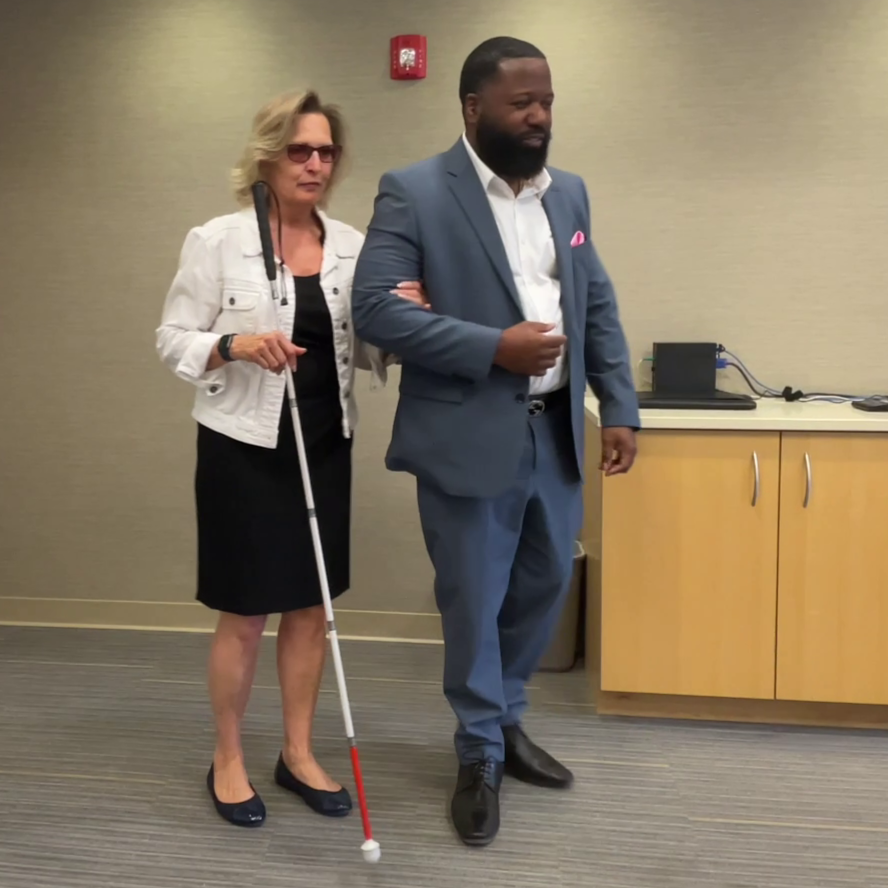 A man uses the human guide technique to guide a woman with a white cane across the room.