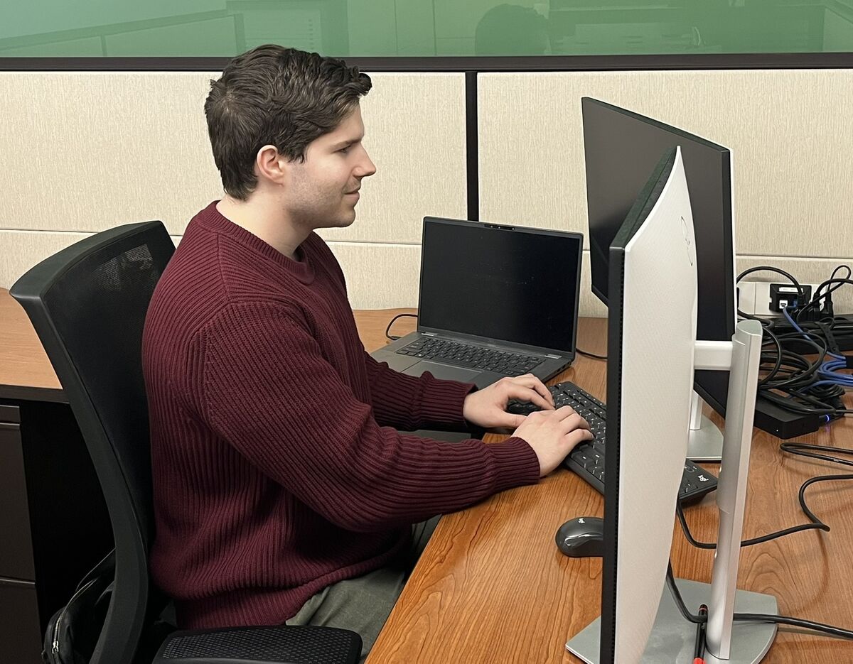 Colin types on a keyboard at his desktop computer in his cubicle. 