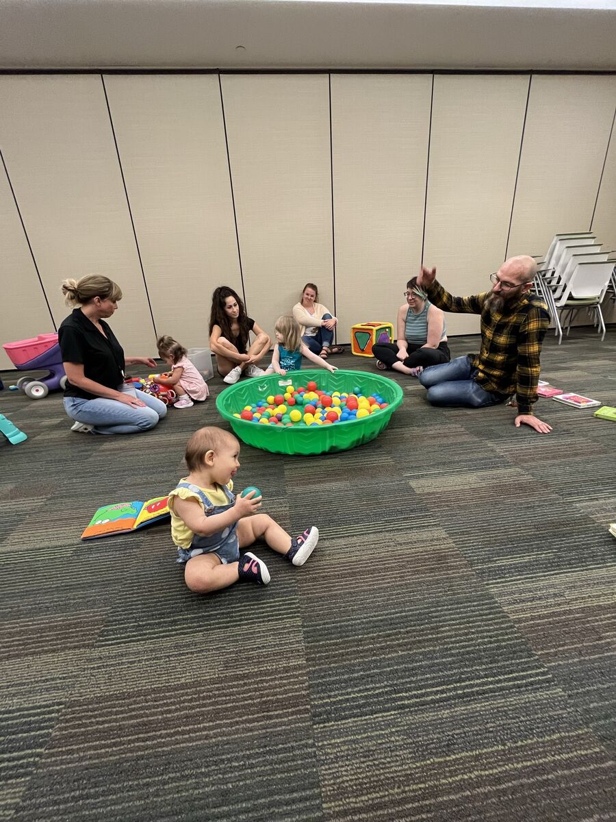 Three parents sit by each other while watching their kids play with toys at CSC's playgroup. Two EI specialists are also watching over the kiddos. 