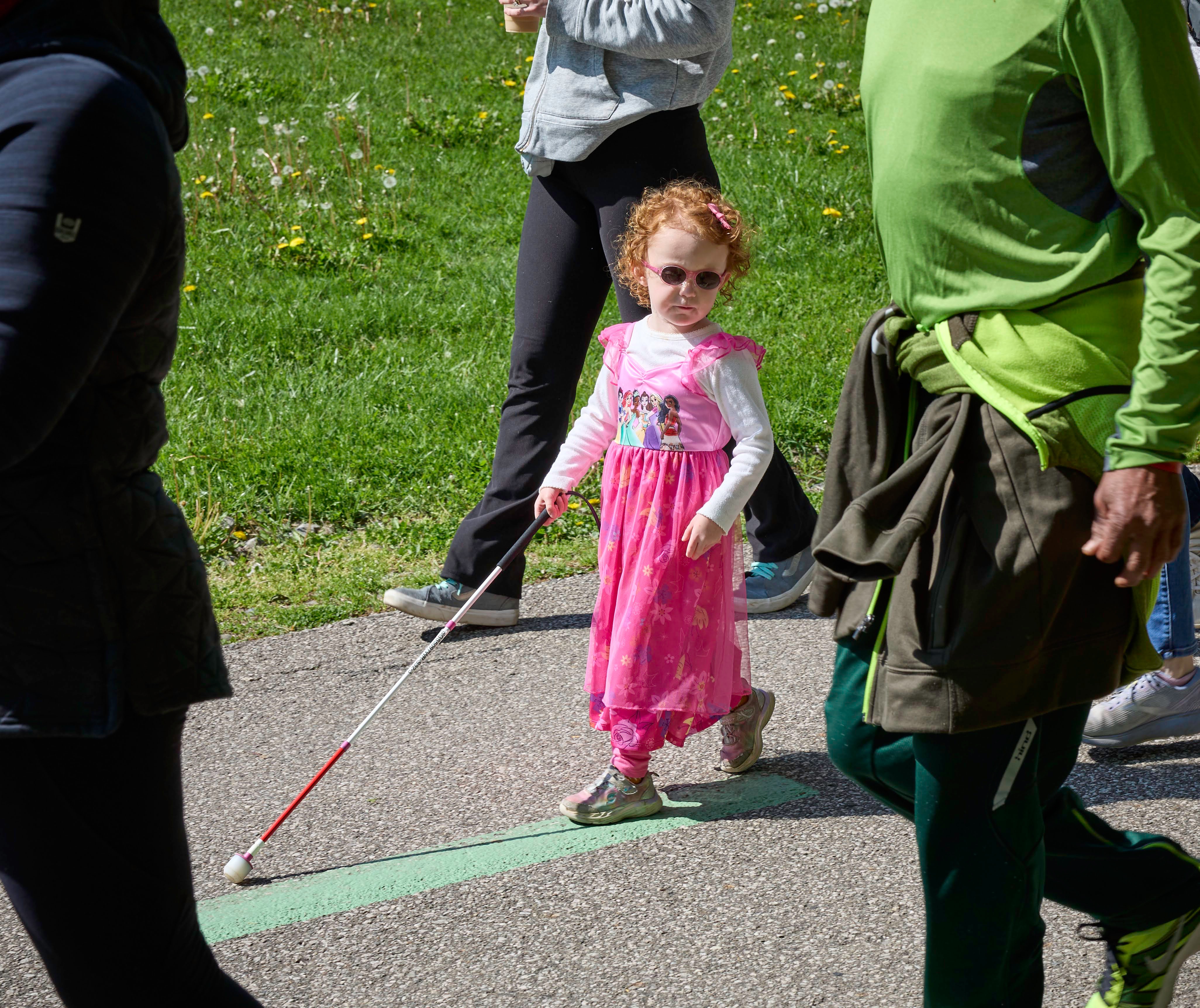 Meet Emma Emma wears a pink princess dress up dress, pink sunglasses and a pink hairbow as she walks on a paved trail.