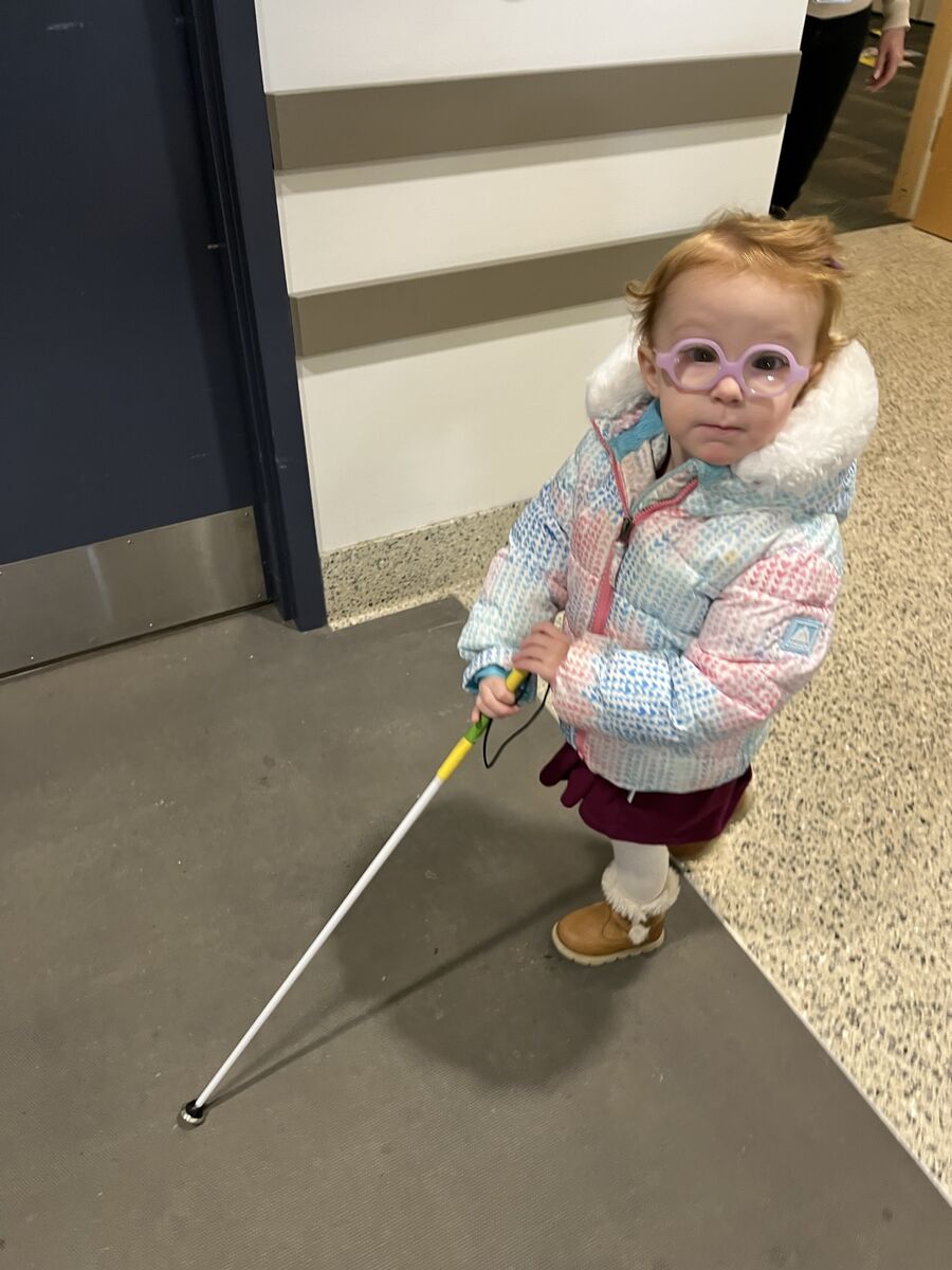A two-year-old Emma looks up at the camera as she walks down a hallway in Cleveland Sight Center using her white cane. She's wearing her big puffy coat for winter weather.