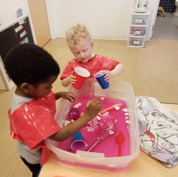 Two preschoolers play with pink colored water while wearing waterproof vests.