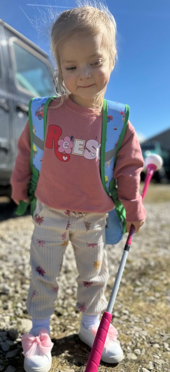 Reese smiles while she's wearing a backpack and holding her white cane. She's wearing a sweatshirt with her name on it and pants with flowers on them.