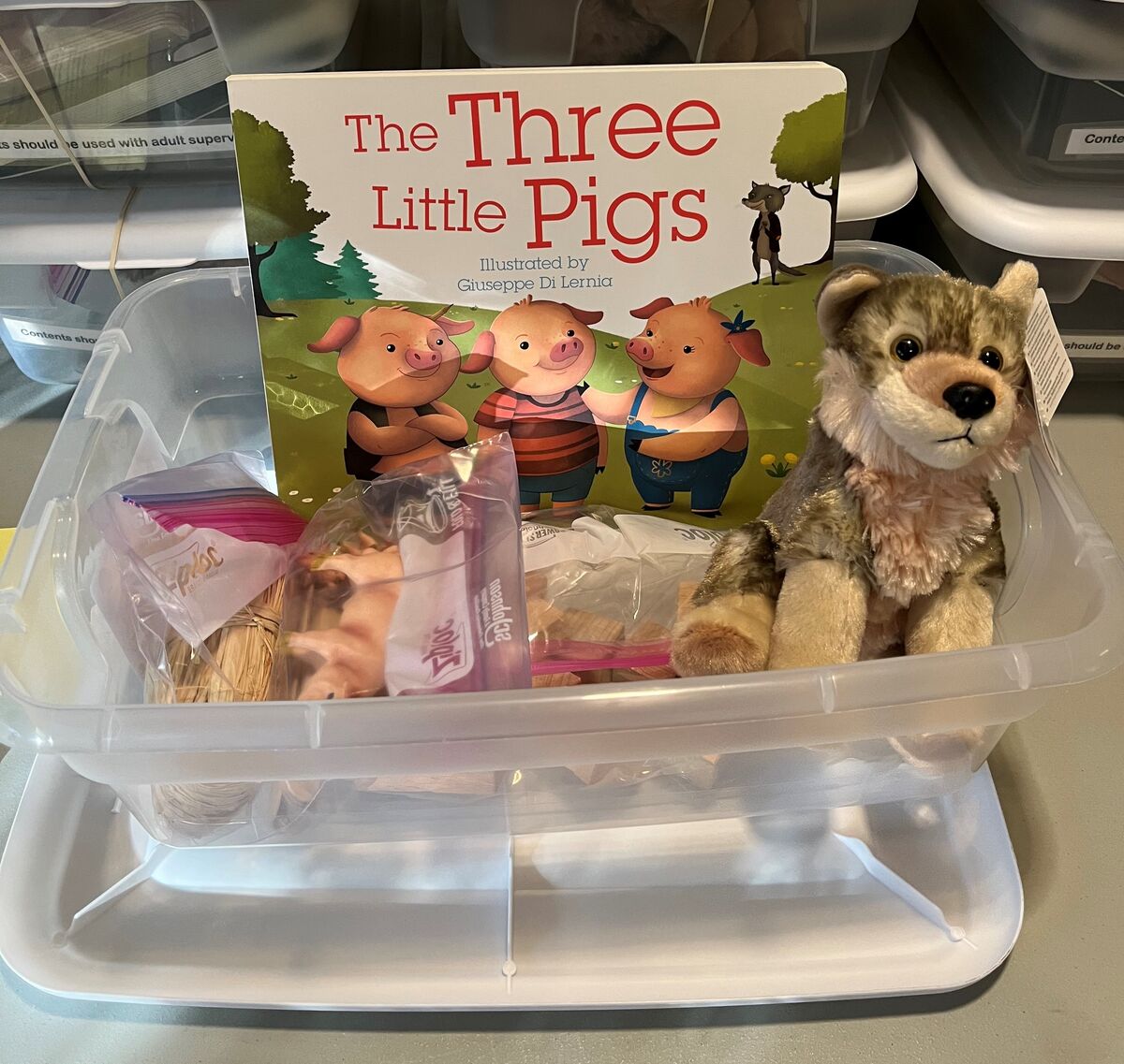 A story box shows the book of the three little pigs, a stuffed animal wolf, pig figurines, bundles of straw and sticks, and a few bricks. 