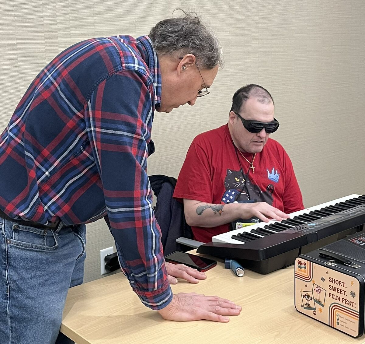 Tom leans over a table with a keyboard piano on it, to supervise a piano class student playing a specific chord.