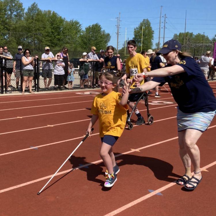 Julianna uses her white cane as she runs on an outdoor track while she holds a helper's hand. She's wearing a yellow shirt and her pink glasses.