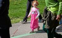 Meet Emma Emma wears a pink princess dress up dress, pink sunglasses and a pink hairbow as she walks on a paved trail.