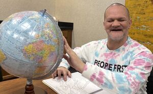 Dale smiles as one hand feels the braille globe in front of him, and another hand reads the braille map on the table in front of him.