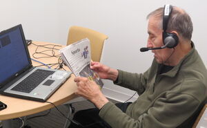 A radio reader reads the sports section of a newspaper aloud into a microphone headset, which is connected to a laptop in front of him.